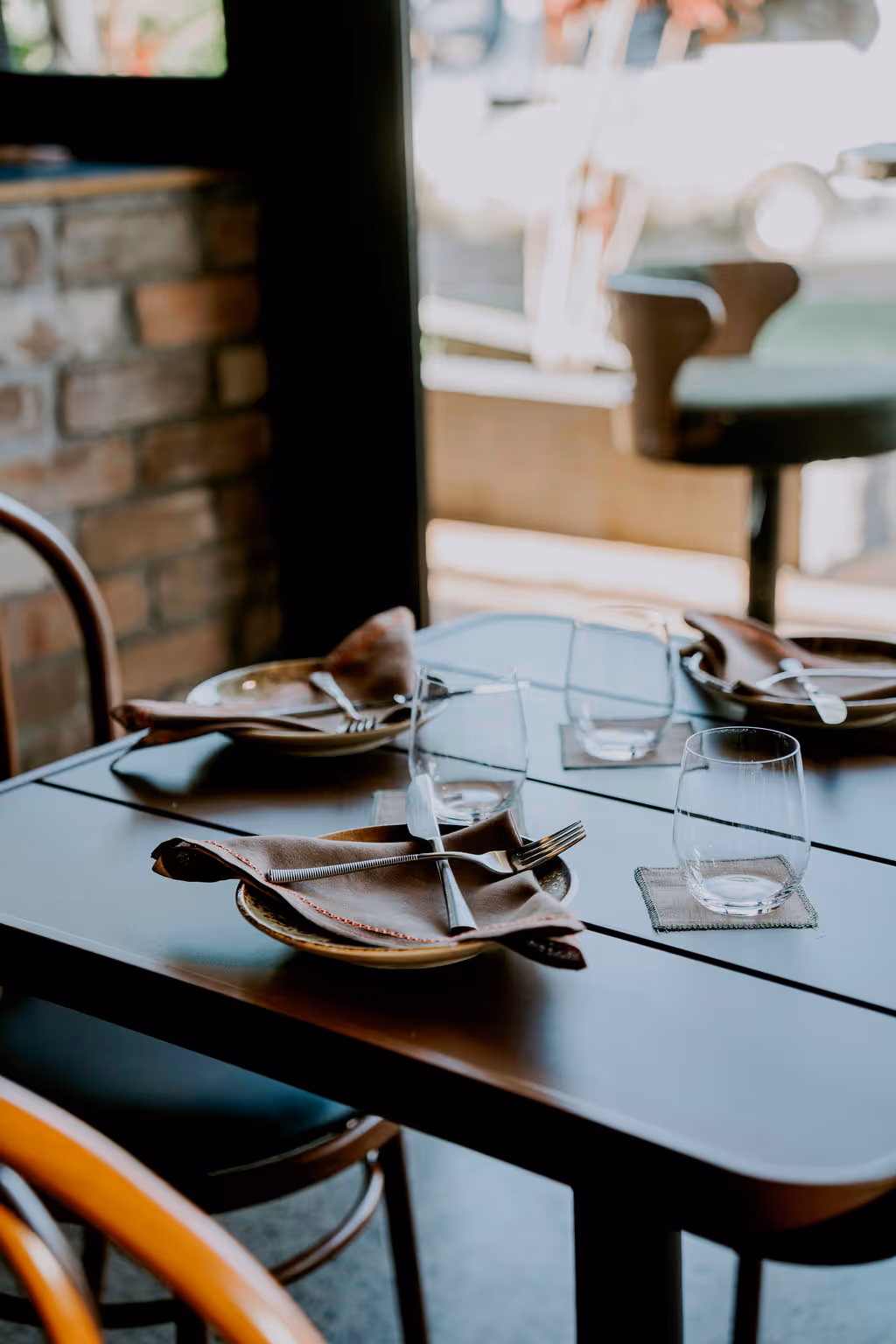 Table set for three with plates, brown napkins, forks, knives, and empty glasses in a restaurant with brick wall and wooden chairs.