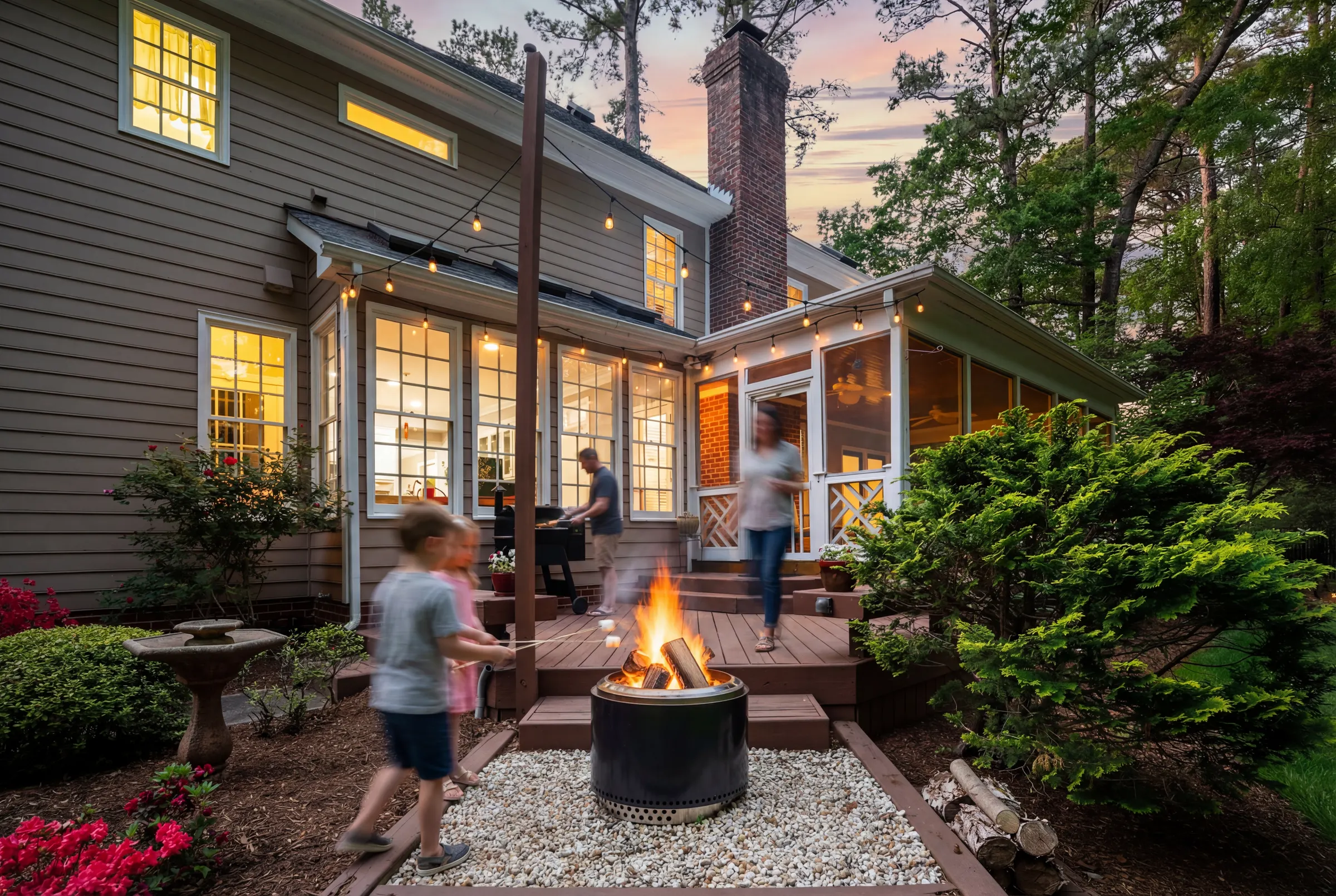 People gathering around a backyard fire pit beside an illuminated home at dusk.