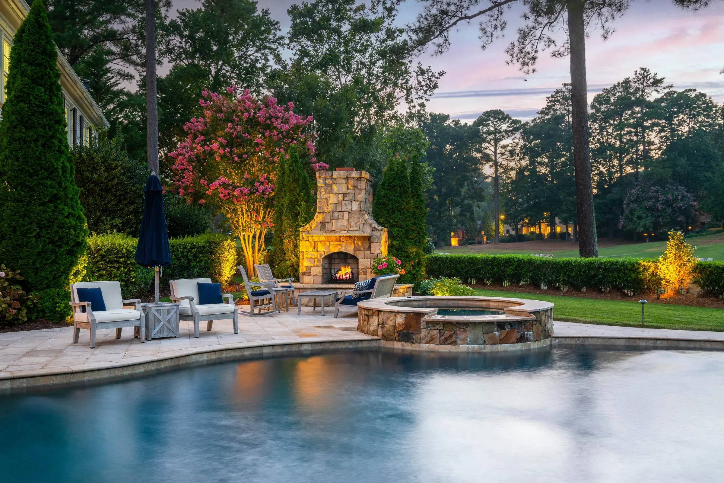 Pool and raised spa beside a stone outdoor fireplace and illuminated landscaping at dusk.