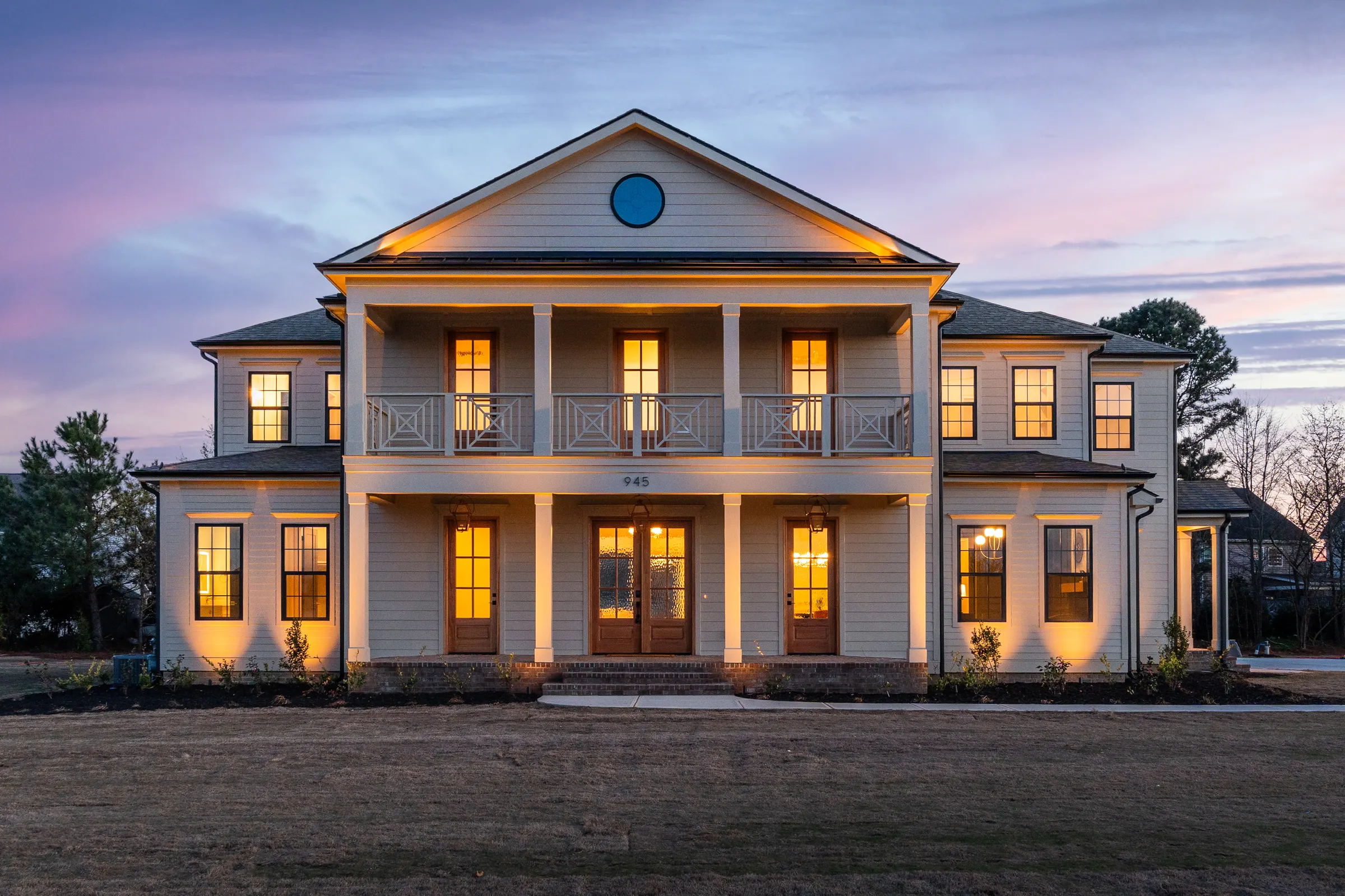 Two-story home with a columned porch, upper balcony, and illuminated windows at dusk.