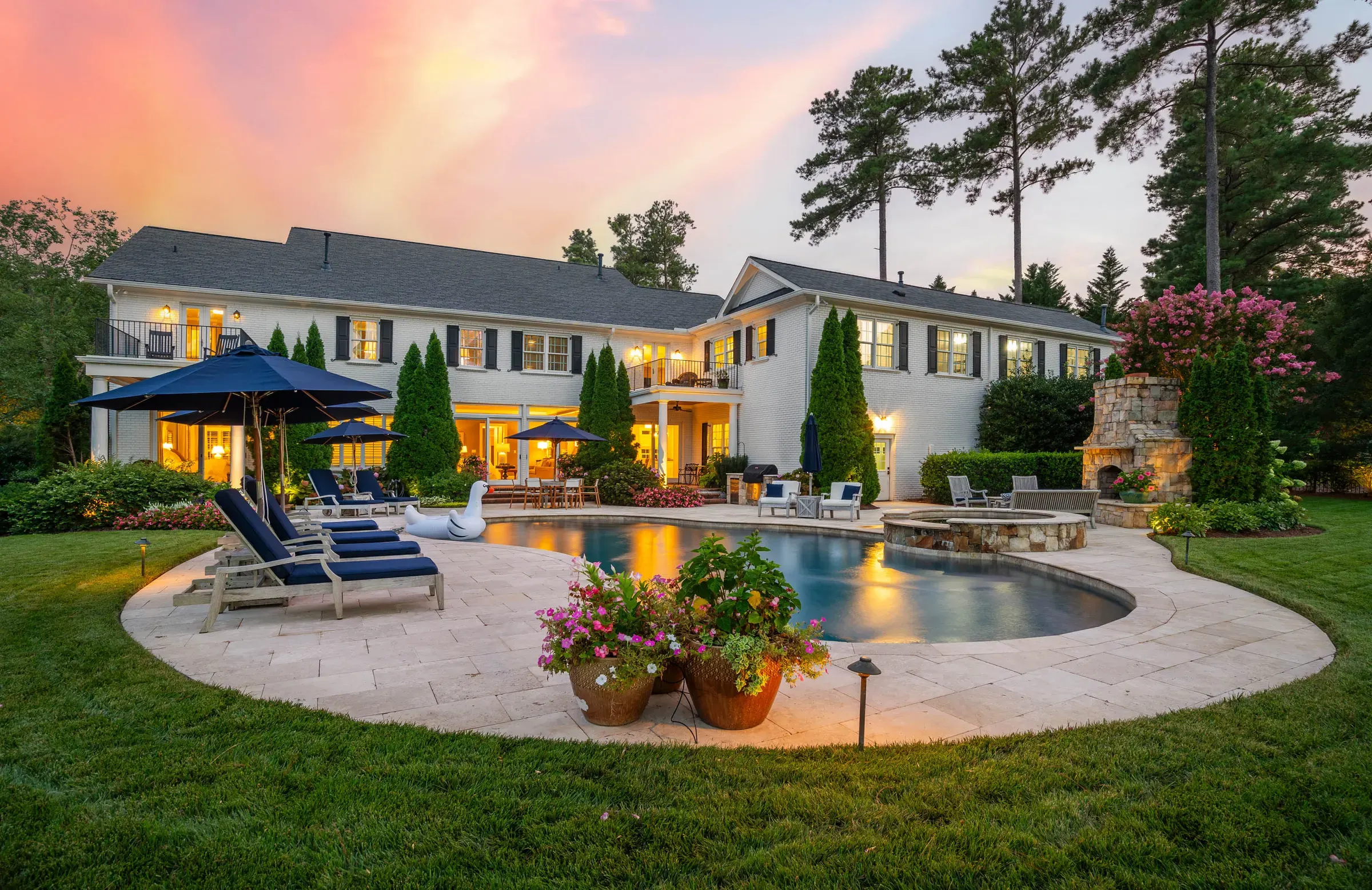 Wide twilight view of a home with a landscaped pool terrace and outdoor fireplace.