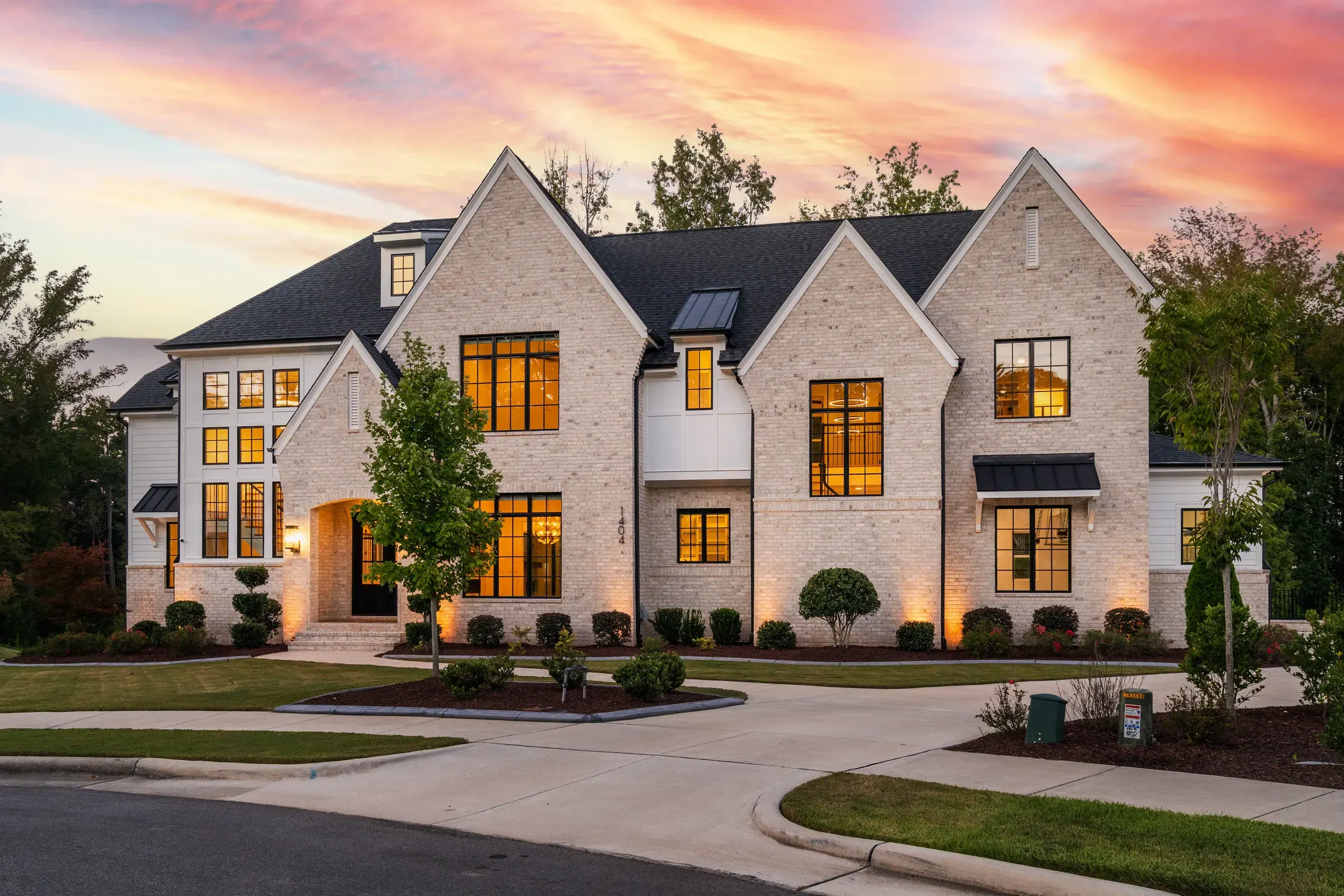 White brick home with glowing windows beneath a pink and orange evening sky.