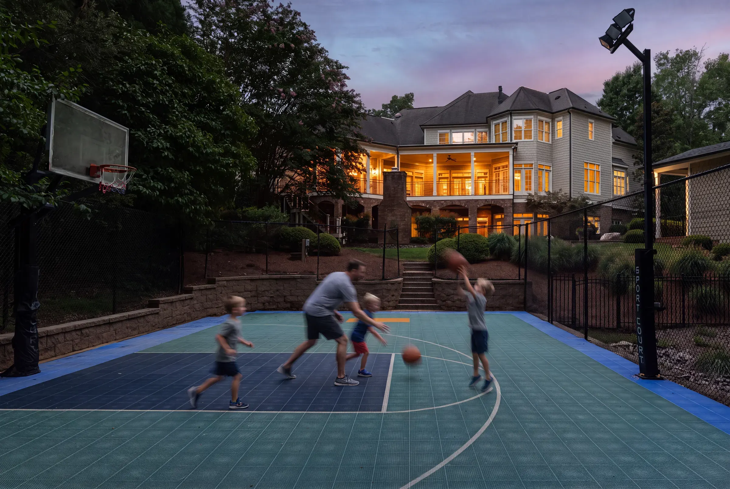 People playing basketball on a backyard court beside an illuminated home at dusk.