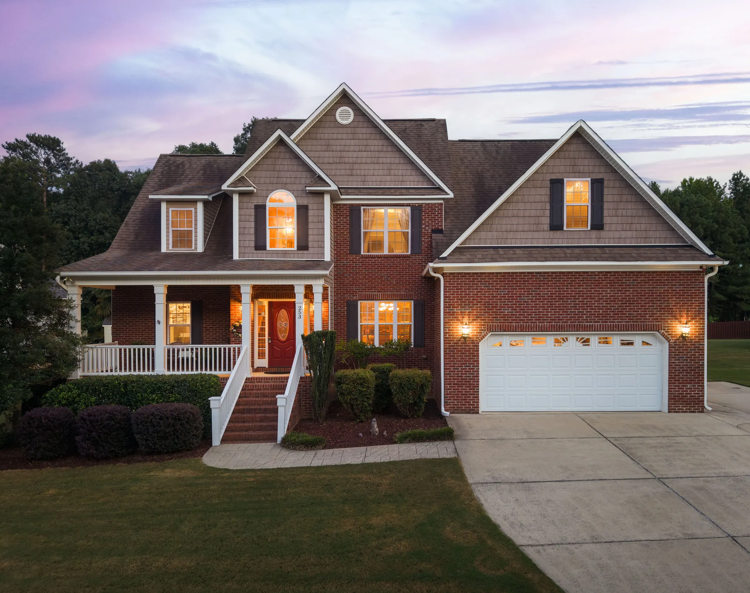 The same brick and shingle home at twilight, with glowing windows and porch lights beneath a pink sky.