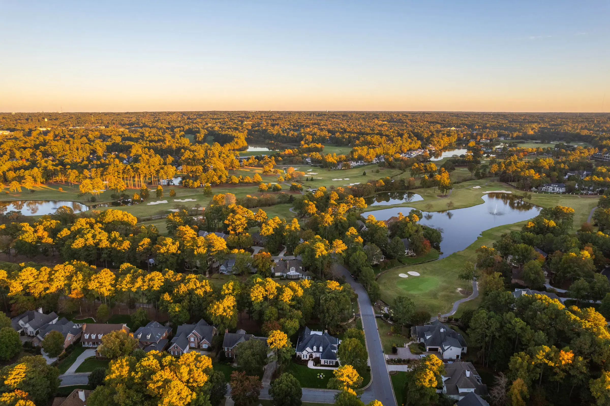 Aerial view of a golf-course community with fairways, lakes, surrounding homes, and wooded landscape.