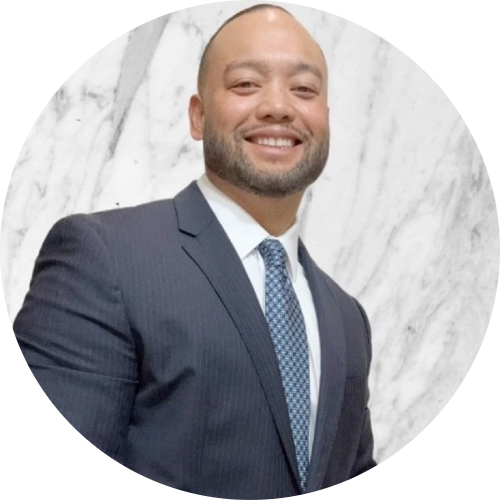 Smiling man in a dark suit and patterned blue tie standing against a white marble background.