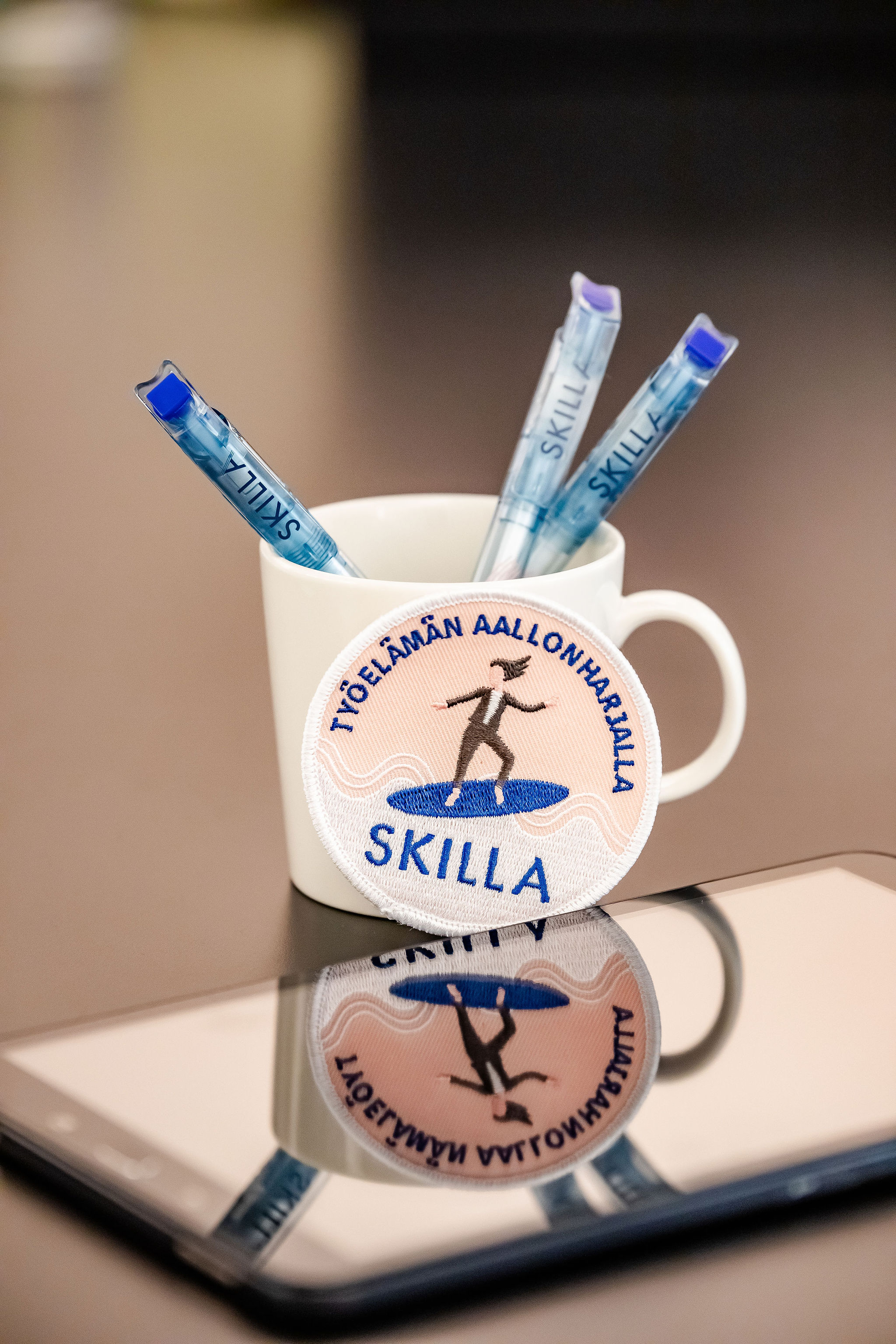 Valkoisessa mukissa on kolme sinistä kynää, ja edessä on Skilla-merkkinen kangasmerkki, joka heijastuu pinnalla olevan tabletin näytössä.