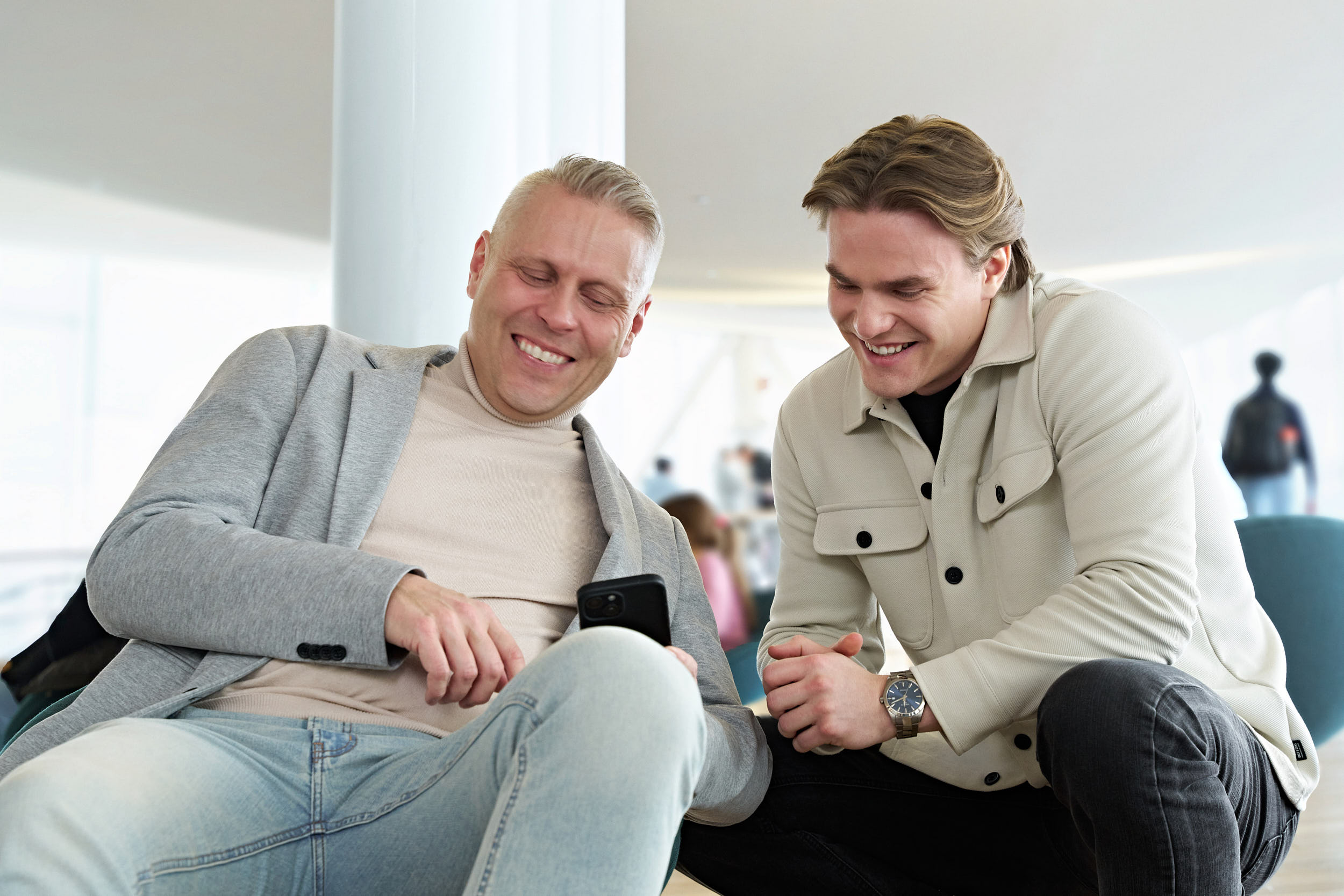 Kaksi miestä istuu ja nojaa eteenpäin katsoen puhelimen näyttöä ja nauraen.