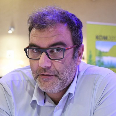 Man with short curly hair, glasses, and a beard wearing a light blue button-up shirt, looking slightly to the side.