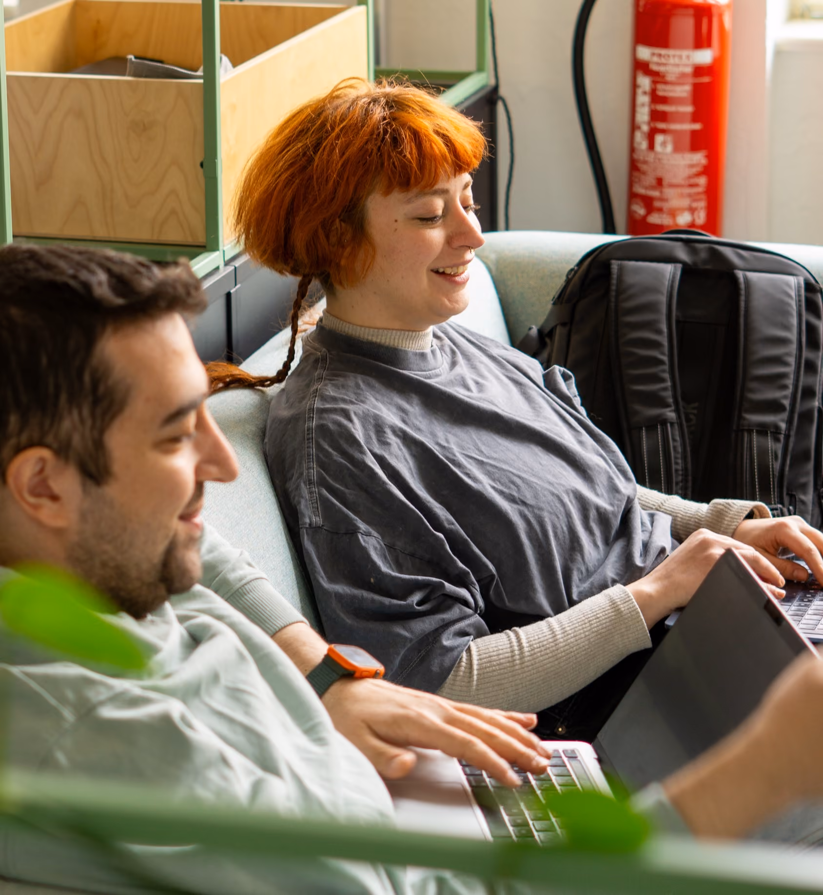 Two young people sitting on a light blue couch working on their laptops together, a red-haired woman smiling and a man with a smartwatch focused on his screen, with a fire extinguisher and backpack in the background.