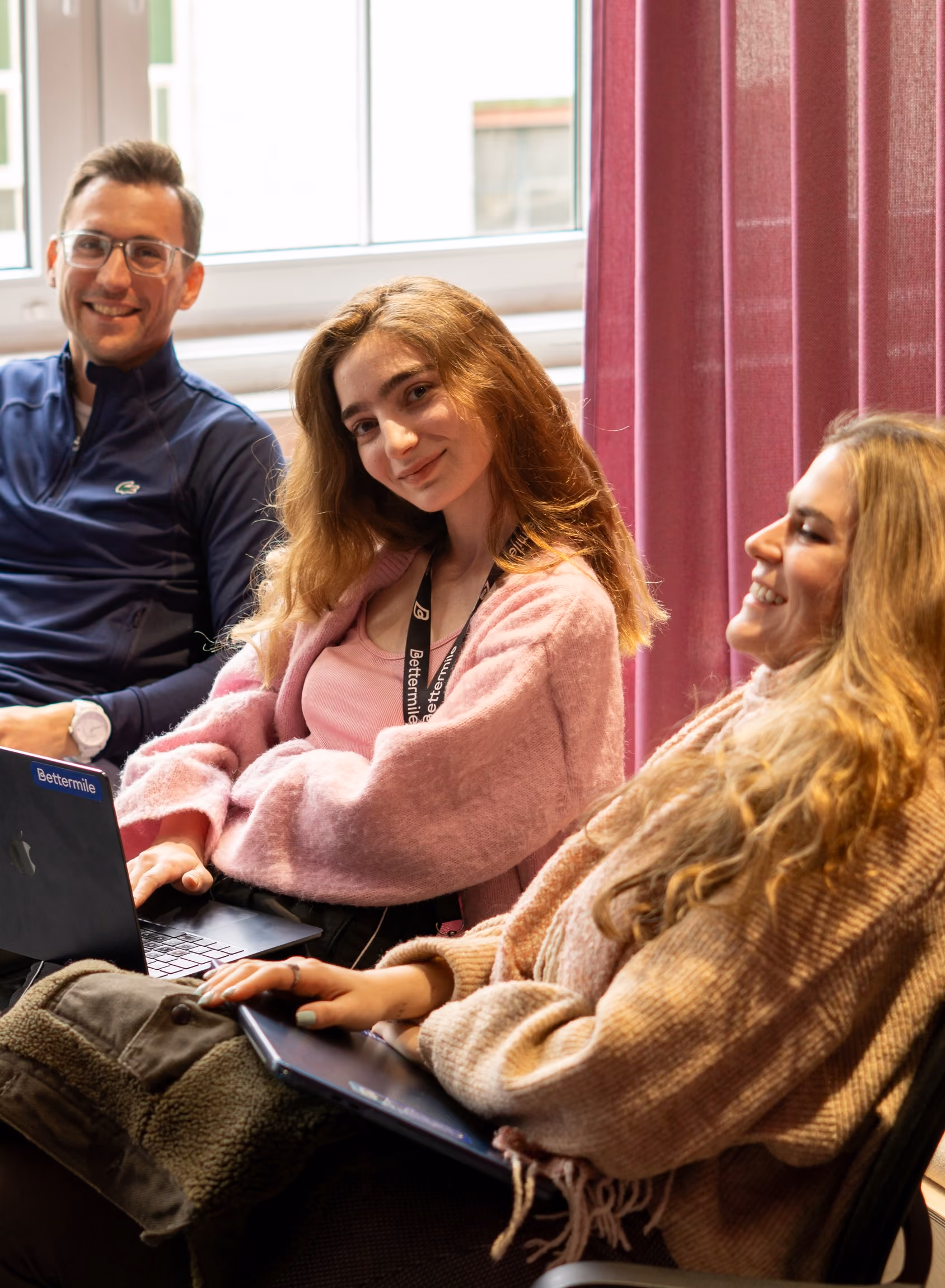 Sales and Marketing Team sitting indoors near a window with a pink curtain, two women smiling and holding laptops, one man in a navy jacket and glasses smiling in the background.