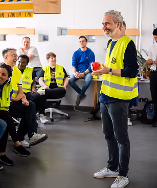 Simon Seeger, Managing Director at Bettermile, in a yellow safety vest speaking with a group of people in safety vests and casual clothes sitting and listening in a bright room.