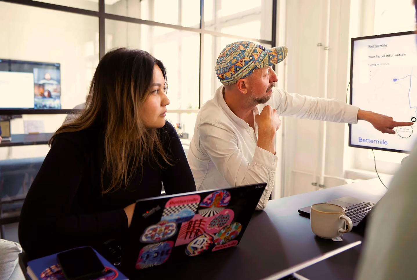 Adrian, Product Owner, wearing a patterned cap points at a screen displaying parcel information and a map while a woman with long hair looks on attentively, both seated at a table with laptops and a coffee cup in a bright office space.