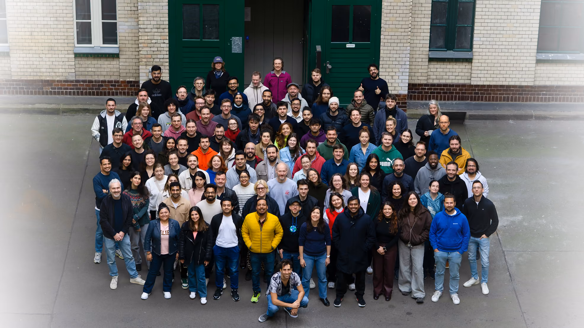 The Bettermile Team: Large diverse group of people posing for a photo together outdoors in front of a building with green doors and beige brick walls.