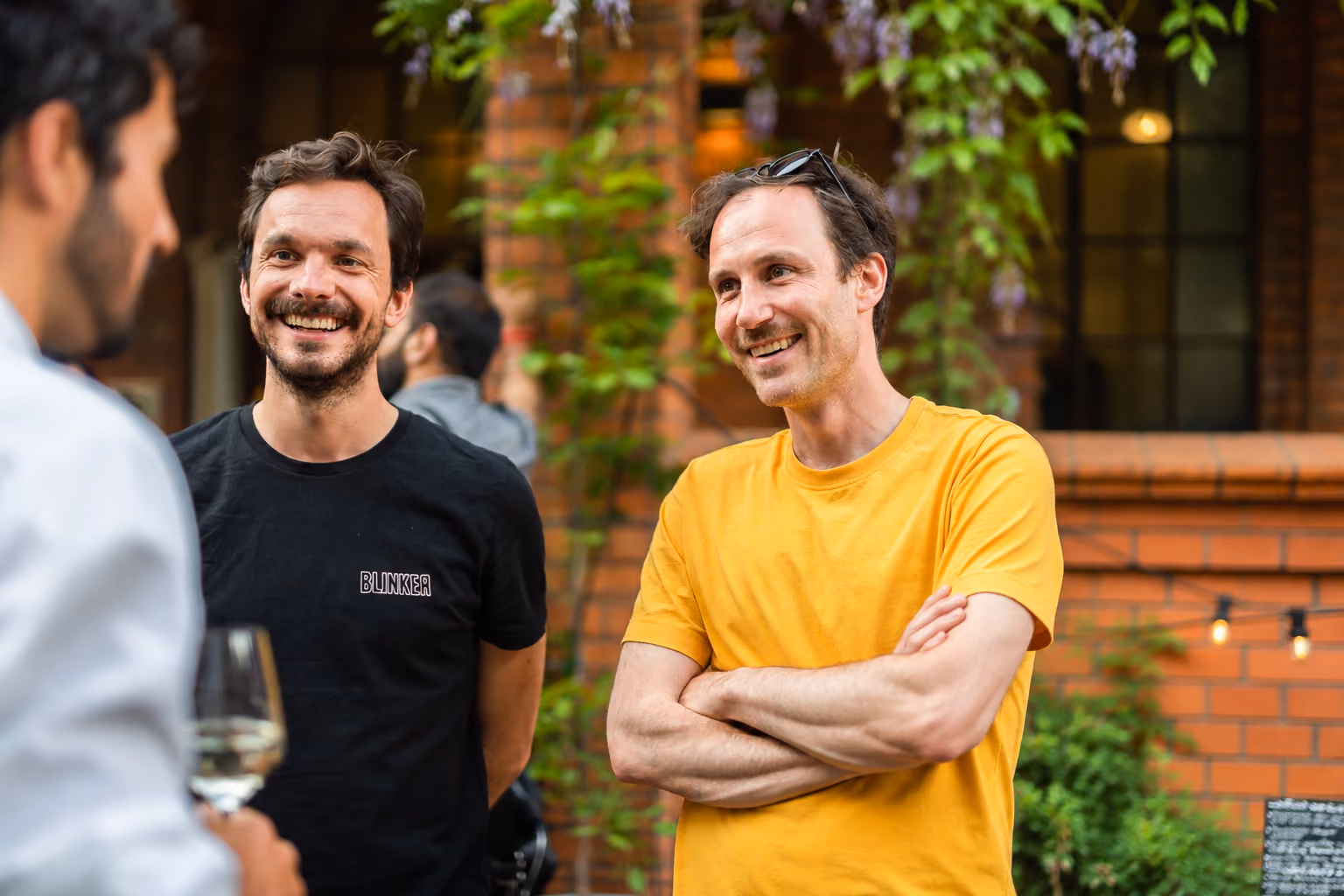 Our Team: Two smiling men engaged in conversation outdoors; the man on the left wears a black T-shirt with 'BLINKER' on it, and the man on the right wears a yellow T-shirt with sunglasses resting on his head. A third person is partially visible on the left holding a wine glass.