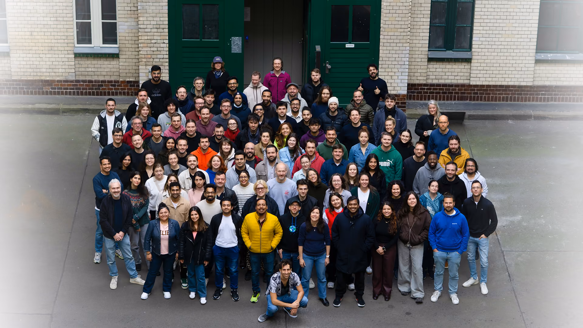 The Bettermile Team: Large diverse group of people posing for a photo together outdoors in front of a building with green doors and beige brick walls.