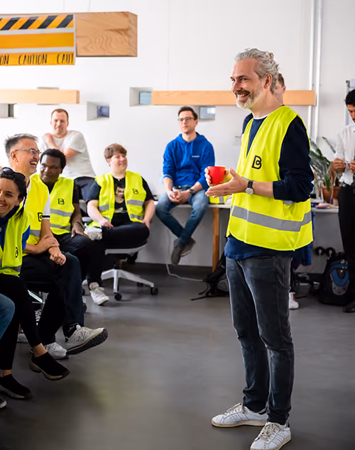 Simon Seeger, Managing Director at Bettermile, in a yellow safety vest speaking with a group of people in safety vests and casual clothes sitting and listening in a bright room.