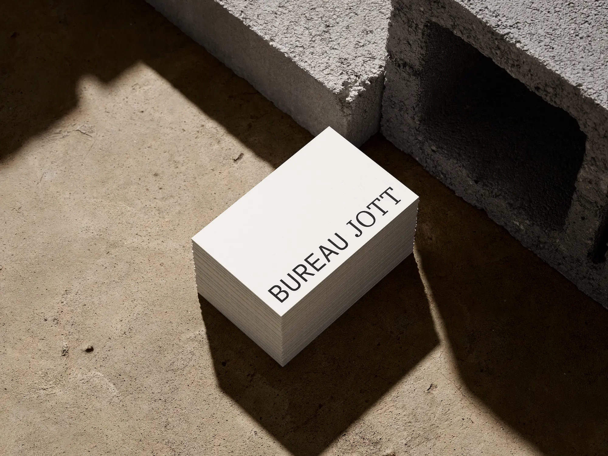 Ein Stapel weißer Visitenkarten mit dem textlichen Aufdruck BUREAU JOTT, liegend auf einem Betonboden neben grauen Betonblöcken.