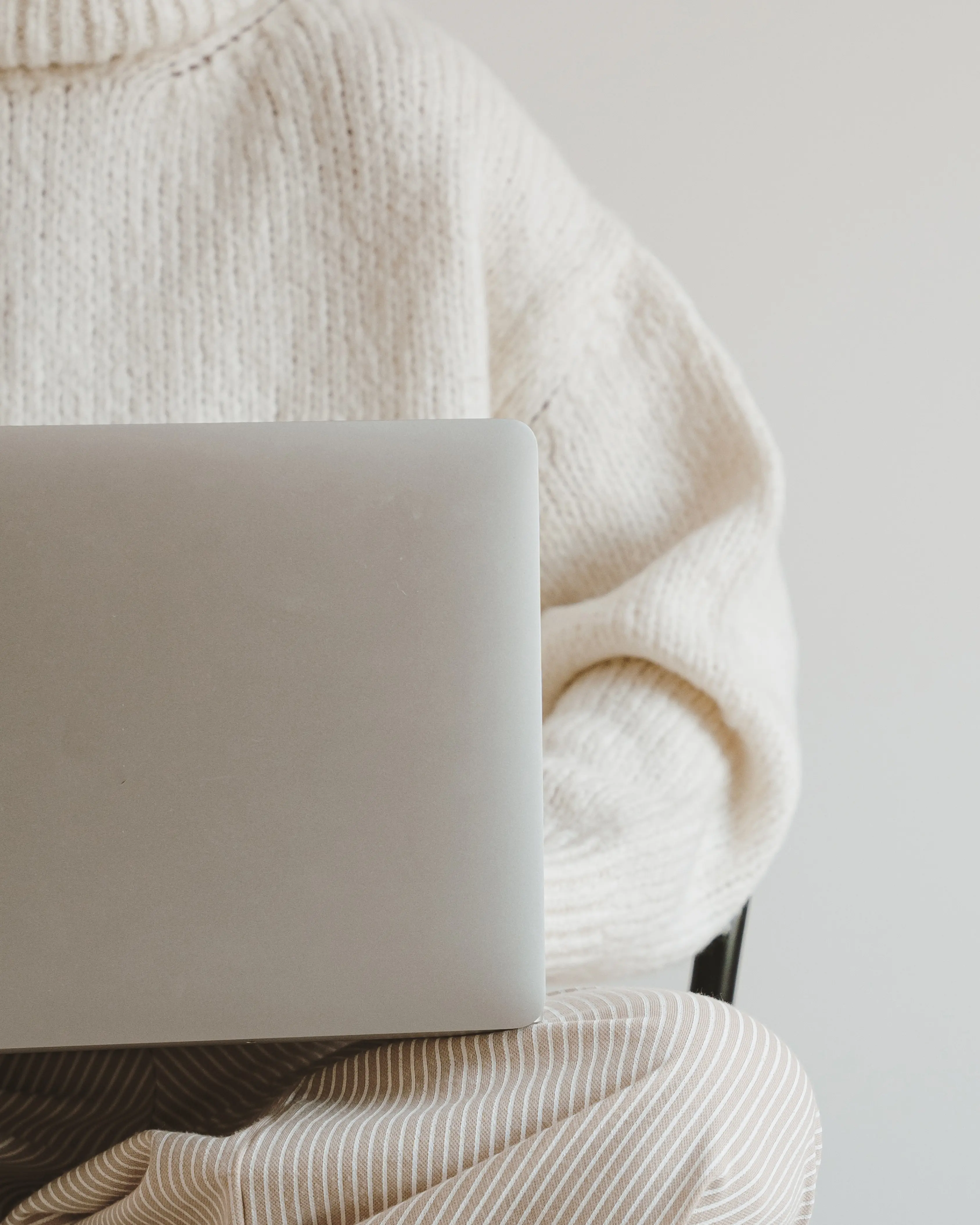 Person mit cremefarbenem Strickpullover und beige-weiß gestreifter Hose, die einen Laptop auf dem Schoß hält.