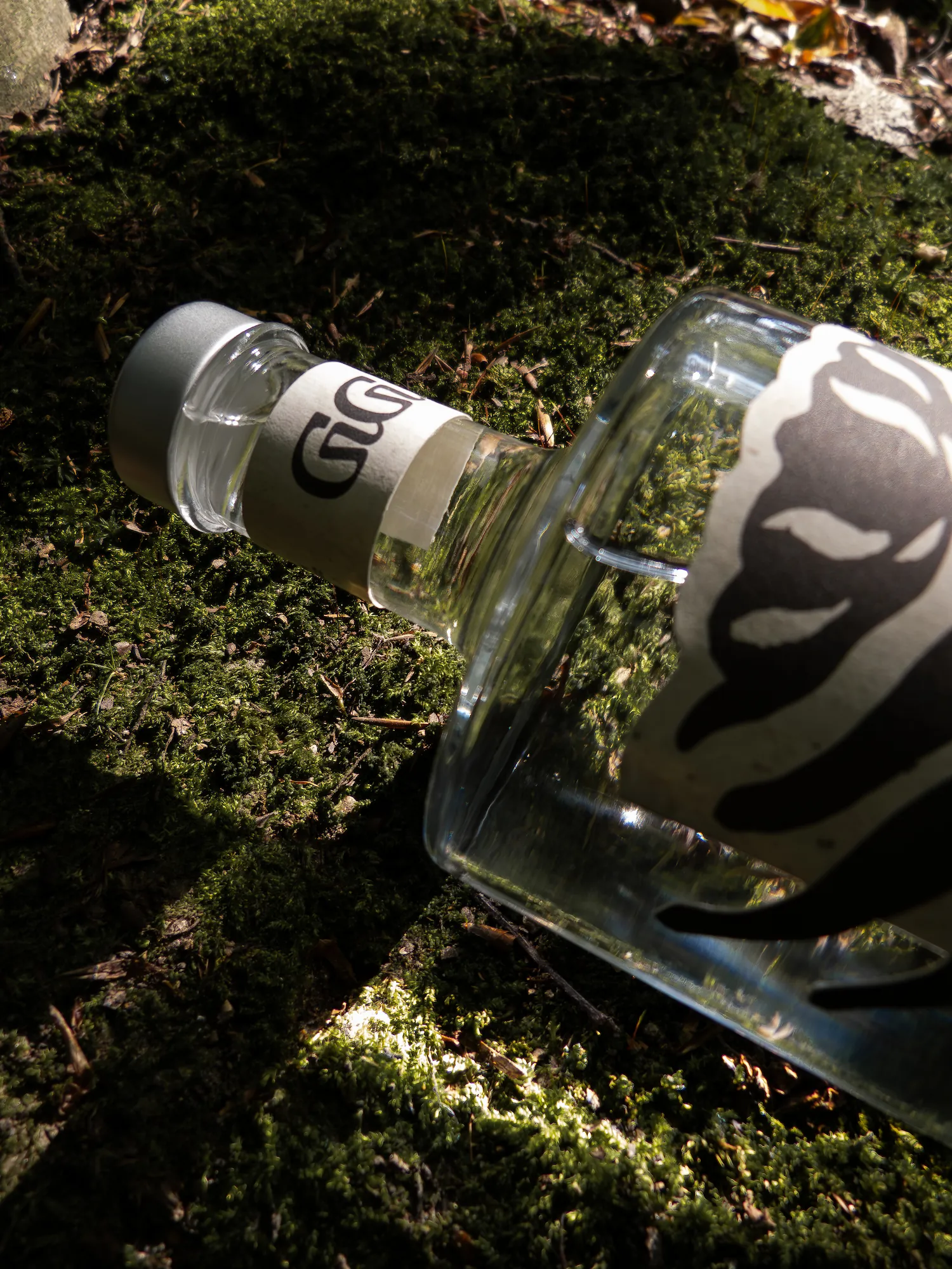 Eine Flasche mit einem silbernen Verschluss und einem Etikett mit schwarzem Text liegt auf moosigem Waldboden.