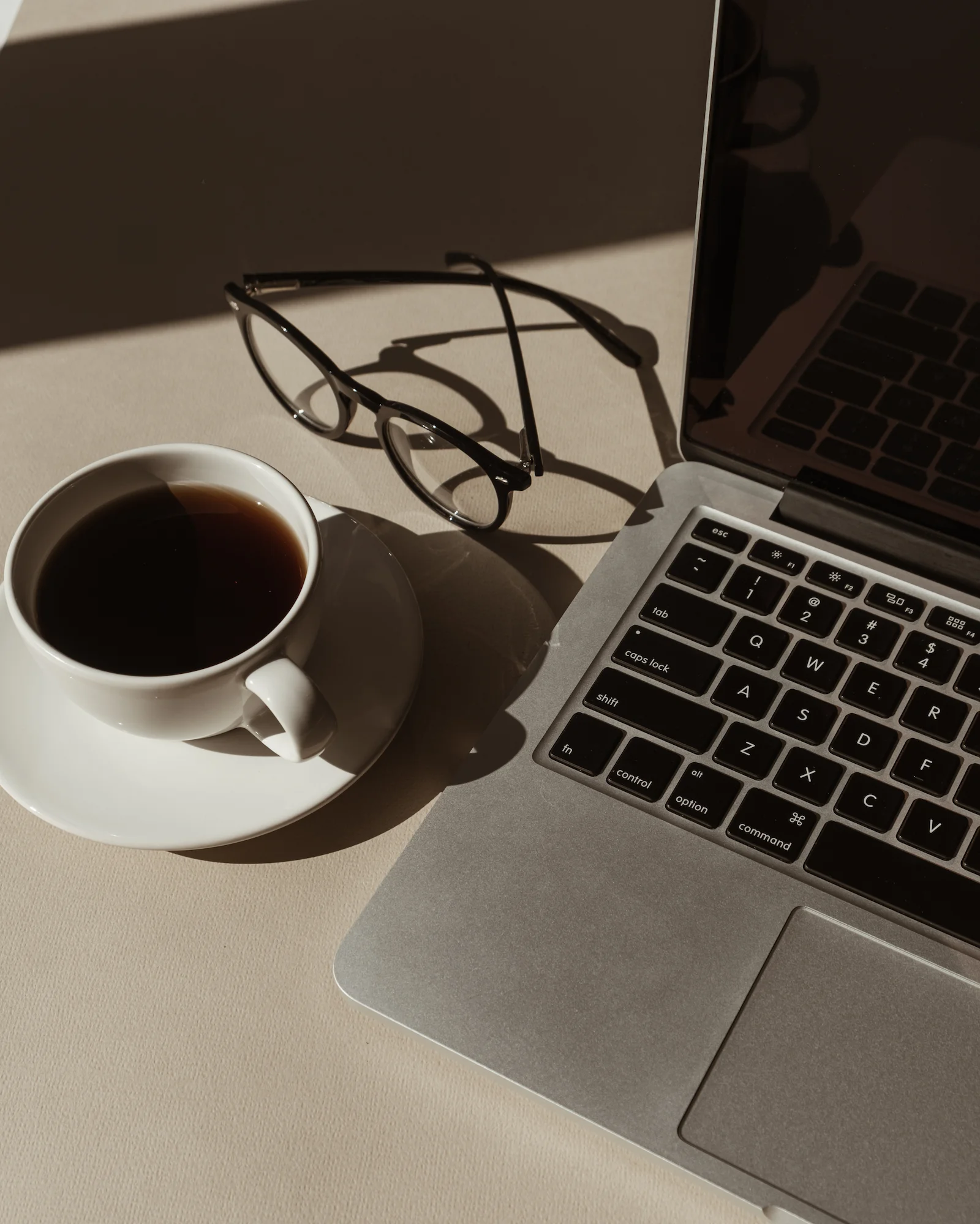 Weiße Tasse mit schwarzem Kaffee, Lesebrille und ein Laptop auf einem hellen Tisch mit Sonnenlicht.