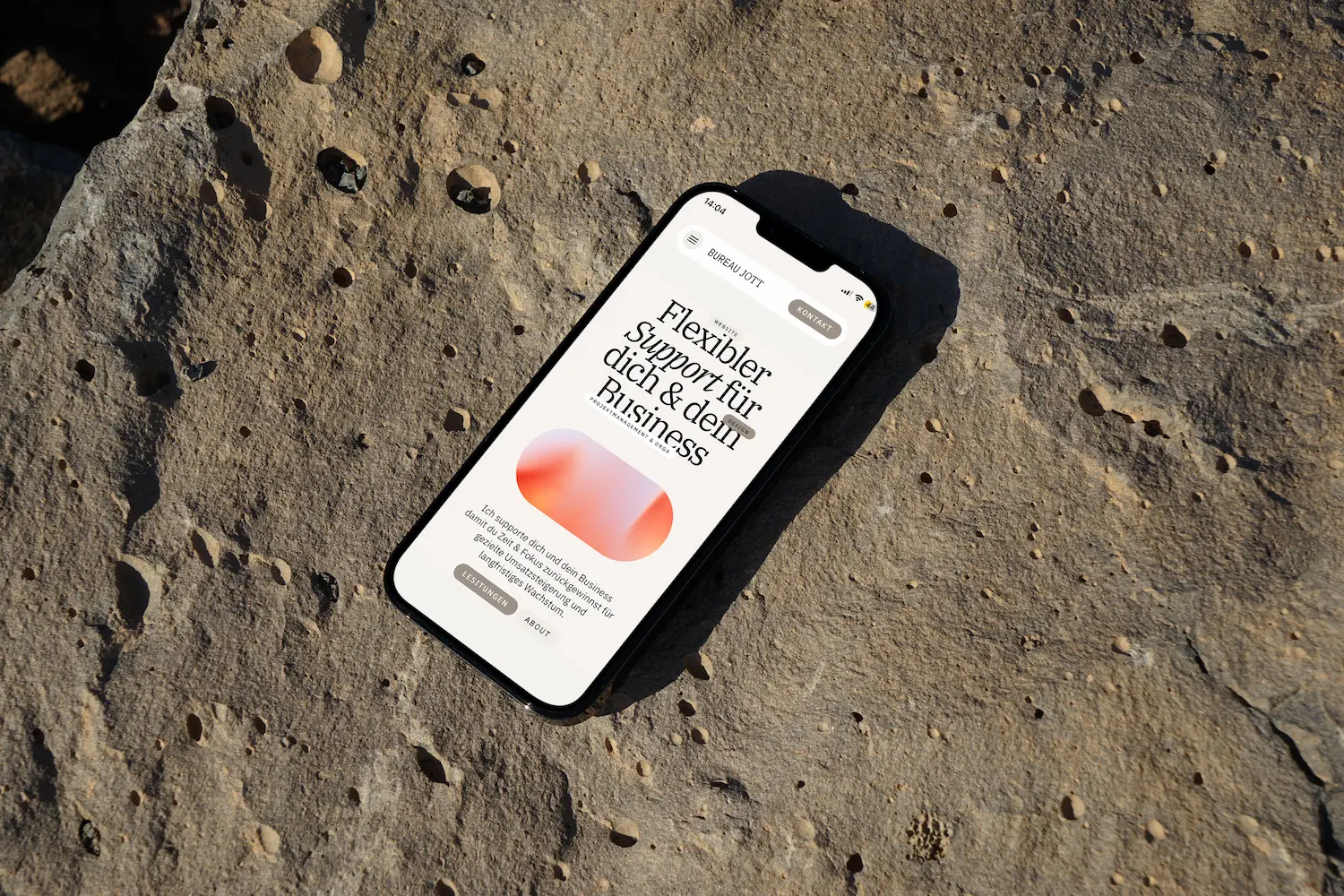 Smartphone auf felsigem Untergrund zeigt eine Website mit dem Text ‚Flexibler Support für dich & dein Business‘ in deutscher Sprache.