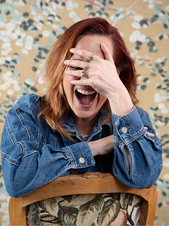 Vrouw met rood haar in spijkerjasje lachend en hand deels voor haar gezicht op een stoel leunend.