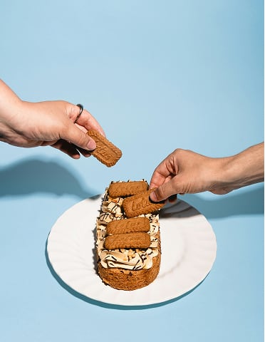 Twee handen plaatsen koekjes op een dessert met room op een wit bord tegen een blauw oppervlak.