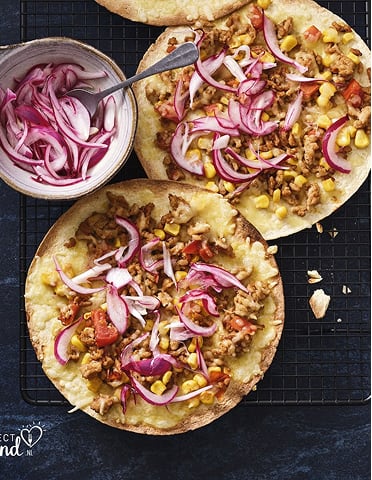 Twee flatbreads met gehakte groenten, maïs en rode ui, naast een kom met extra rode ui.