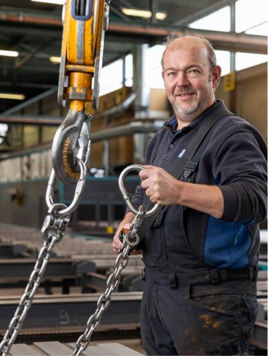 Man met overall houdt ketting vast die is bevestigd aan een hijshaak in een industriële werkplaats.
