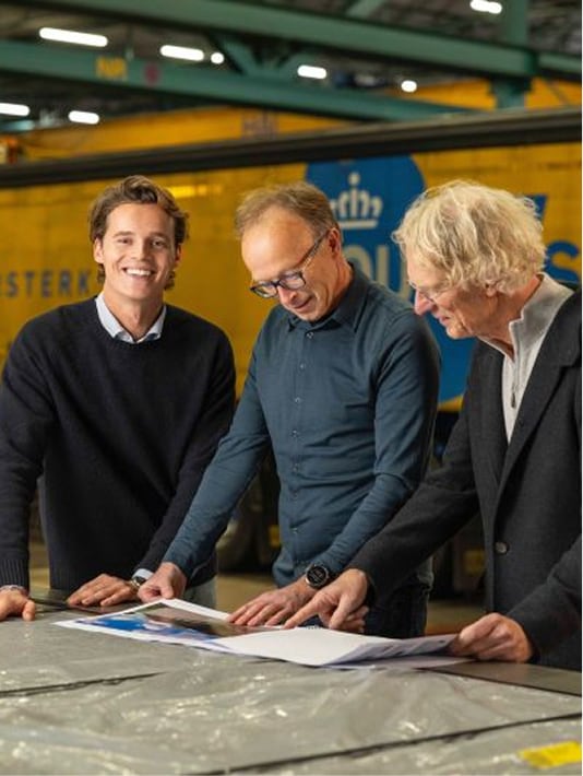 Drie mannen bekijken documenten op een werkbank in een industriële omgeving met geel blauwe achtergrond.