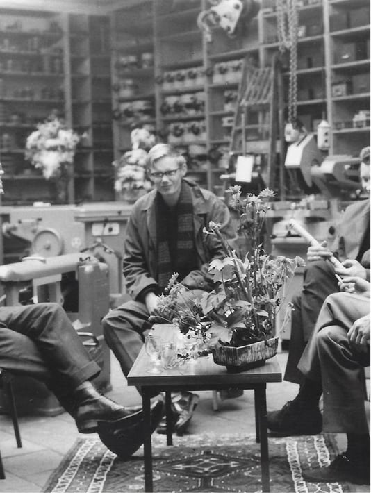 Mensen zitten rond een tafel met bloemen in een werkplaats met machines en gereedschap.