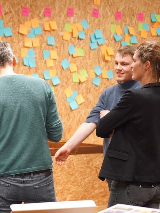 Drie mensen bespreken ideeën bij een muur vol met kleurrijke plakbriefjes.