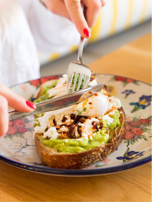 Hand snijdt toast met avocado, geitenkaas, gepocheerd ei en balsamico op een bloemenbord.