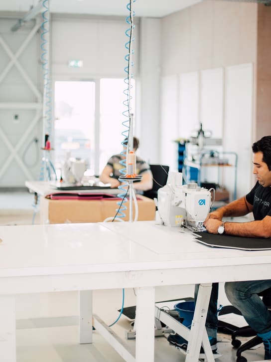 Man working on a sewing machine in a bright workshop with fabric and another person in the background.