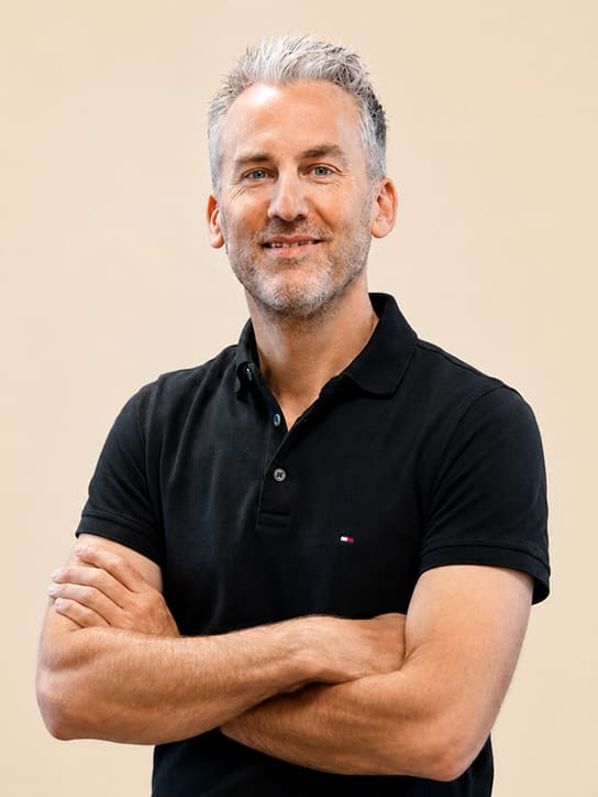 Man met grijs haar en baard lacht terwijl hij zijn armen over elkaar heeft in een zwarte polo.
