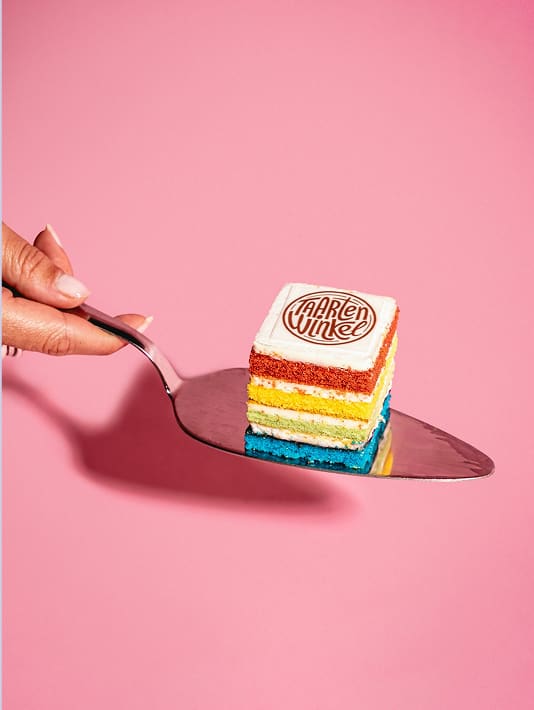 Kleine laagjestaart met regenboogkleuren op een taartspaan tegen een roze achtergrond.