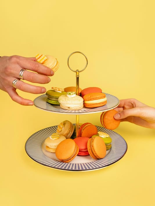 Twee handen pakken kleurrijke macarons van een tweelaags bord tegen een gele achtergrond.