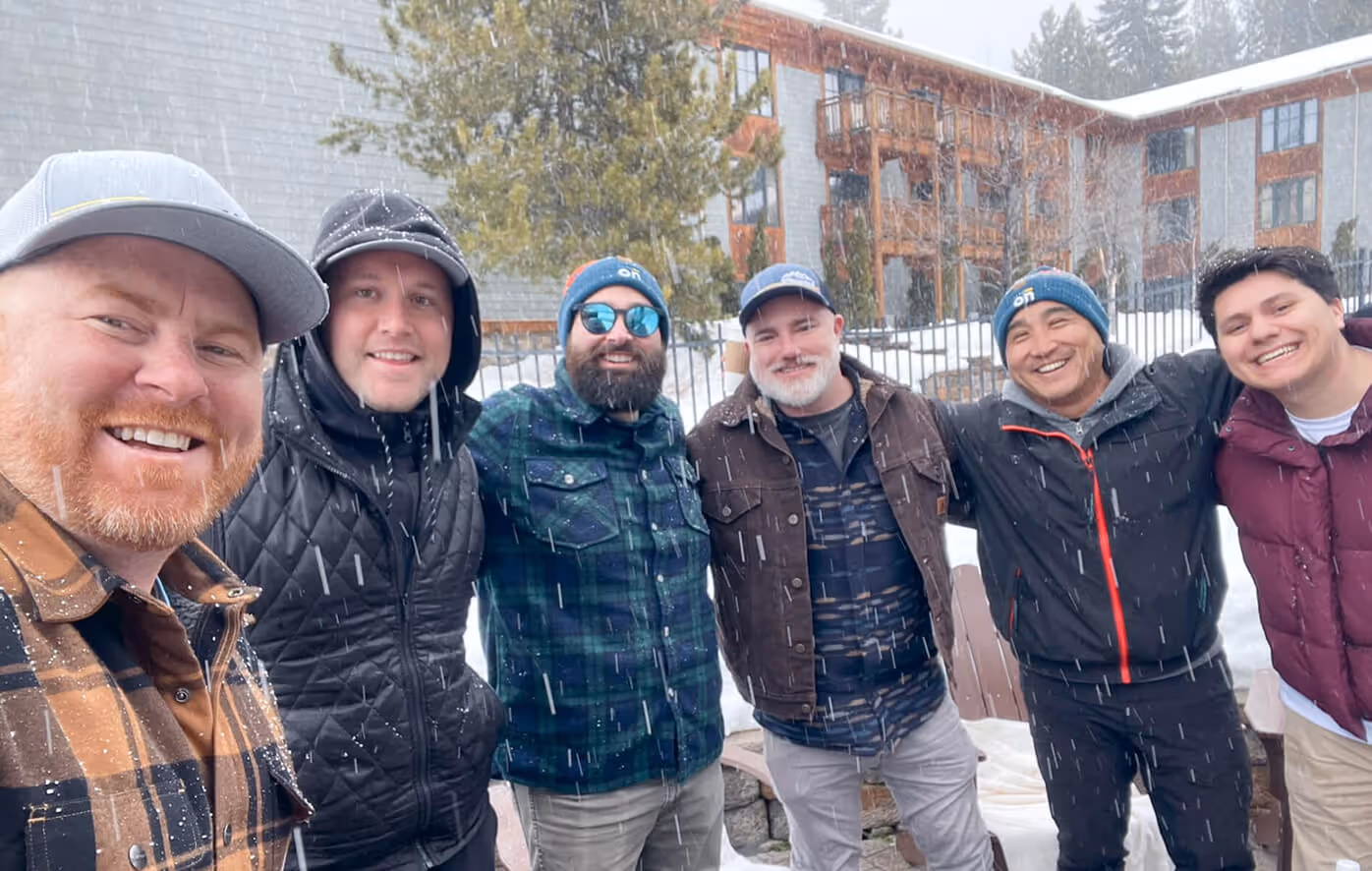 Six men smiling and posing together outdoors in light snow near a building with balconies.