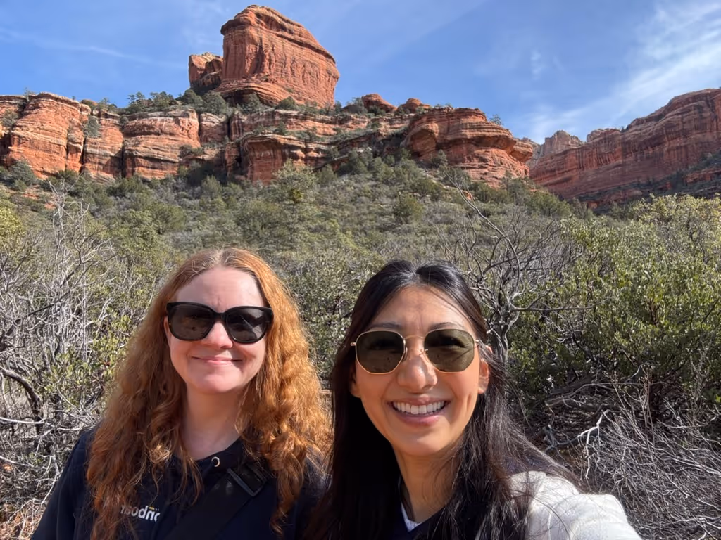 Two women wearing sunglasses smiling with red rock formations and green vegetation in the background under a blue sky.