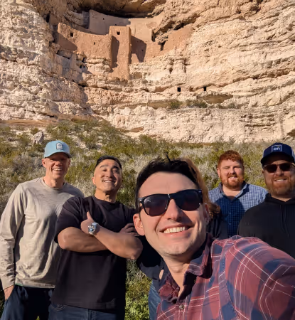 Five men smiling for a selfie outdoors with ancient cliff dwellings visible in the rock formation behind them.