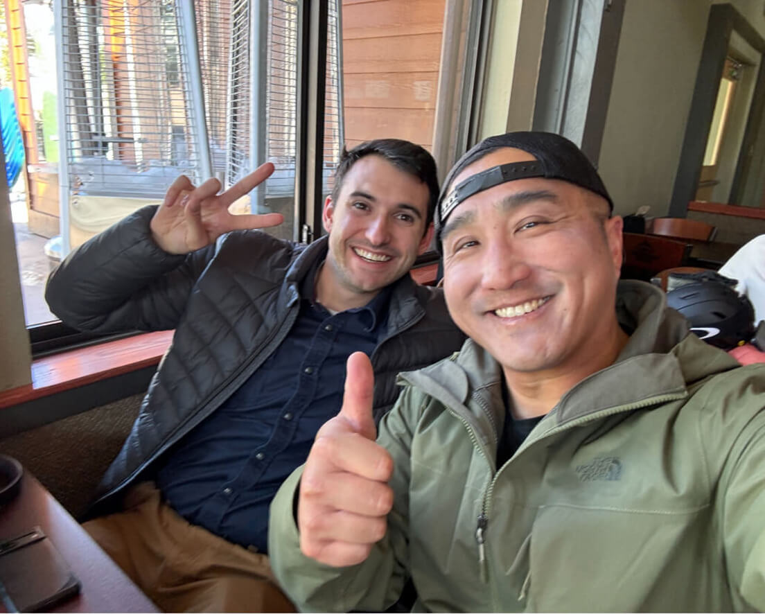 Two men indoors smiling at the camera, one giving a thumbs up and the other making a peace sign.