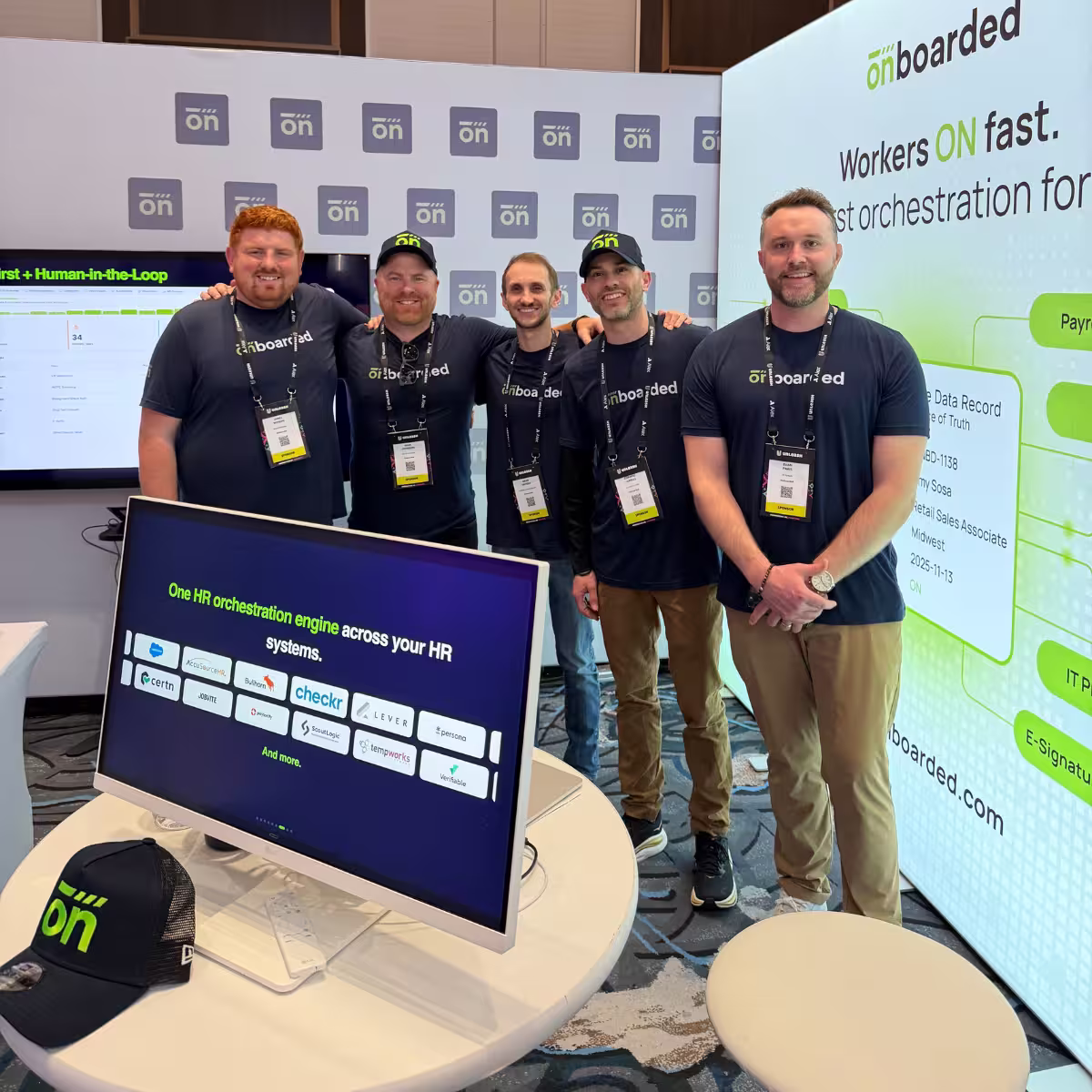 Five men wearing matching Onboarded t-shirts and lanyards smiling and standing together at a conference booth with Onboarded branding and a monitor displaying HR orchestration information.