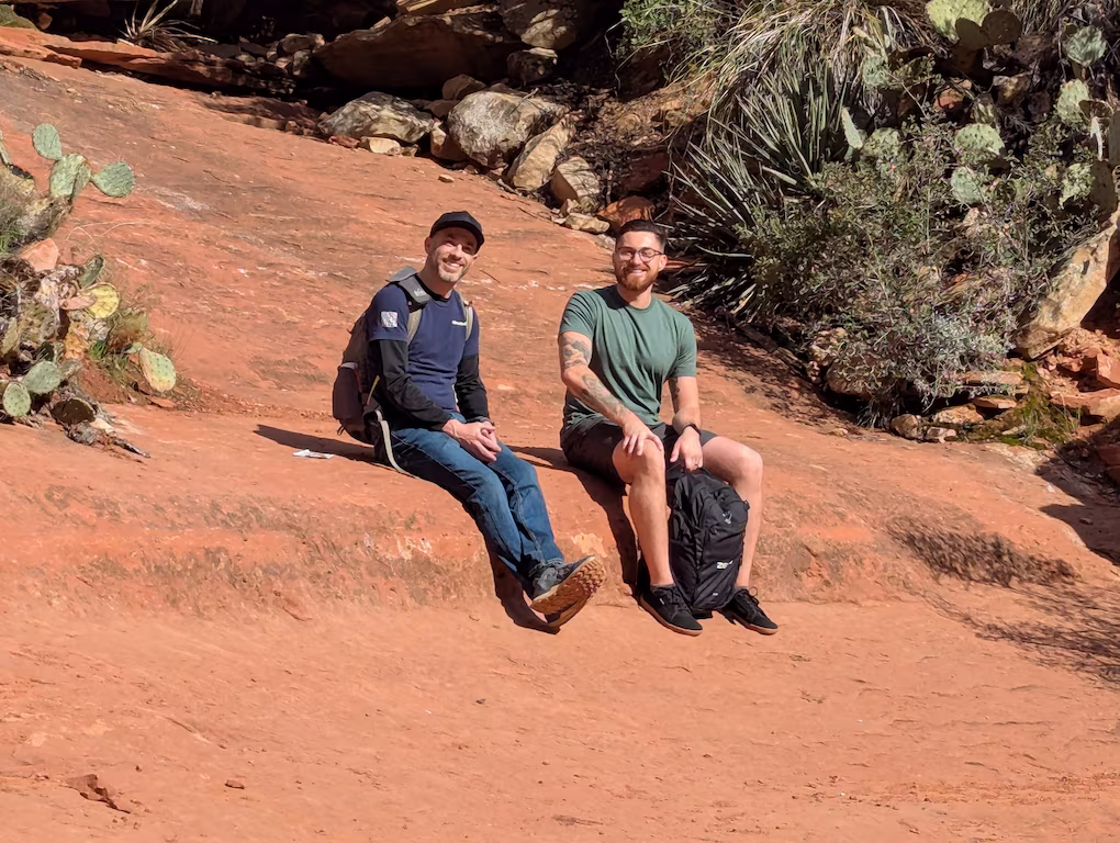 Two men sitting on a red rock surface with backpacks, surrounded by desert vegetation including cactus and shrubs.
