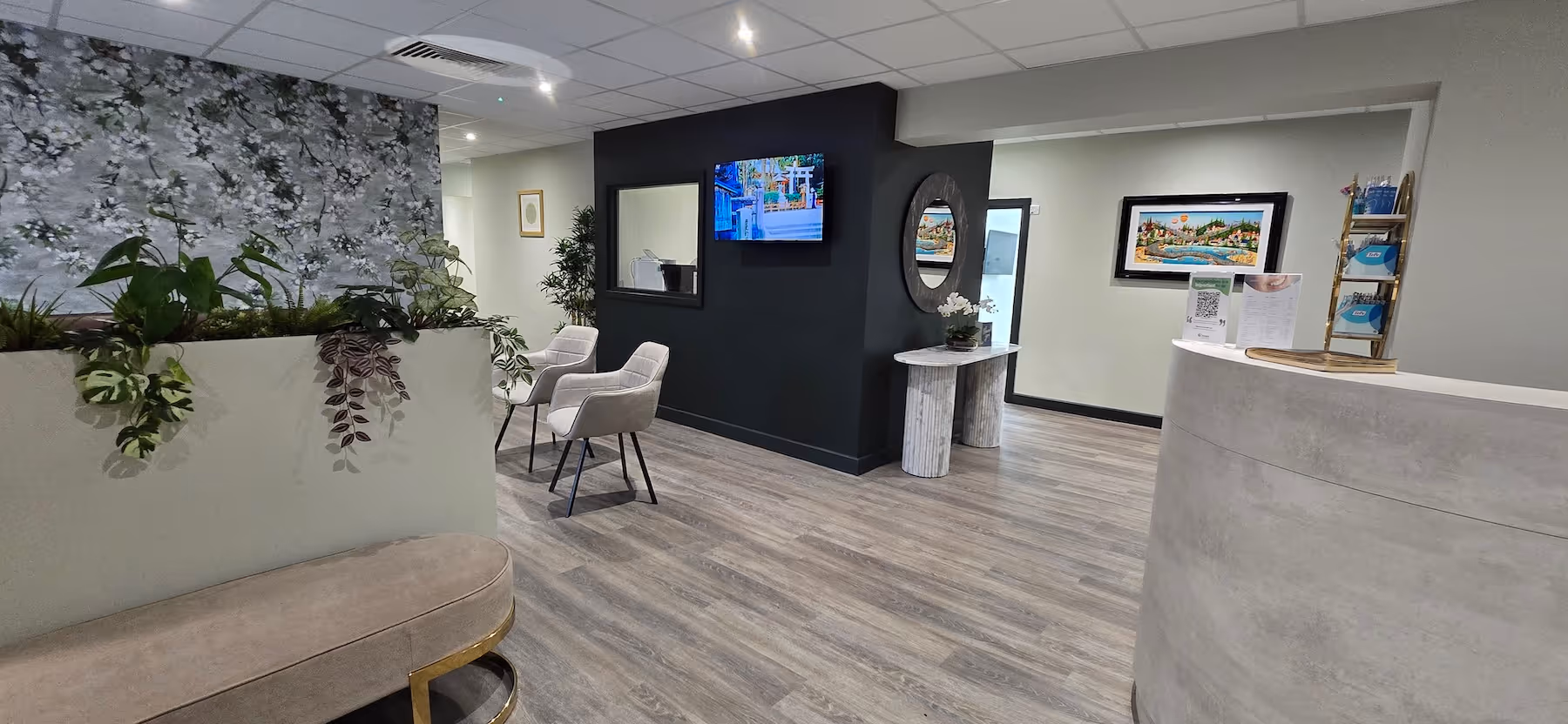 Modern reception area with light wood flooring, gray armchairs, a floral partition, a curved reception desk, wall-mounted TV, and decorative wall art.