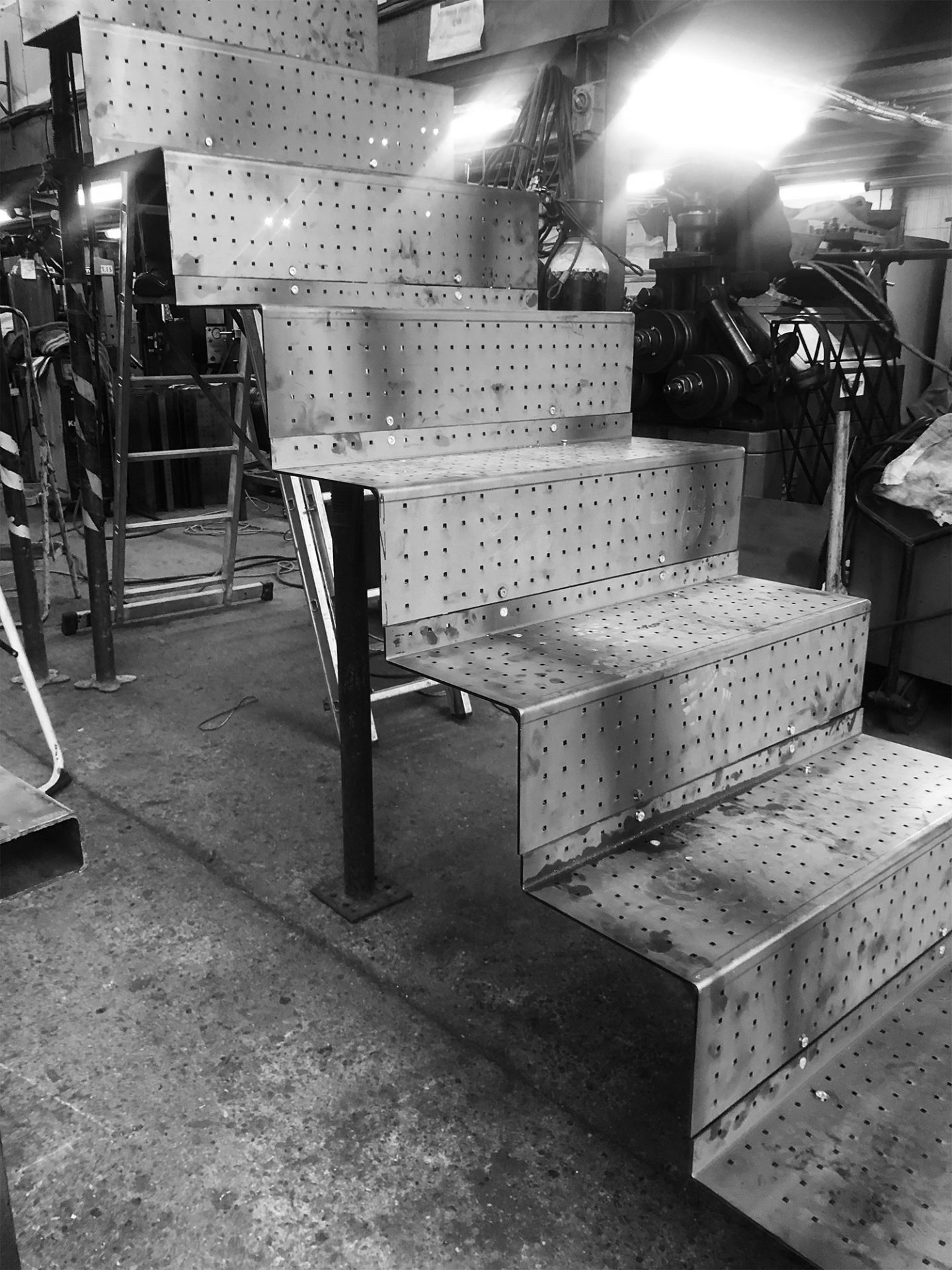 Metal staircase under fabrication in an industrial workshop with perforated steps and welding marks.