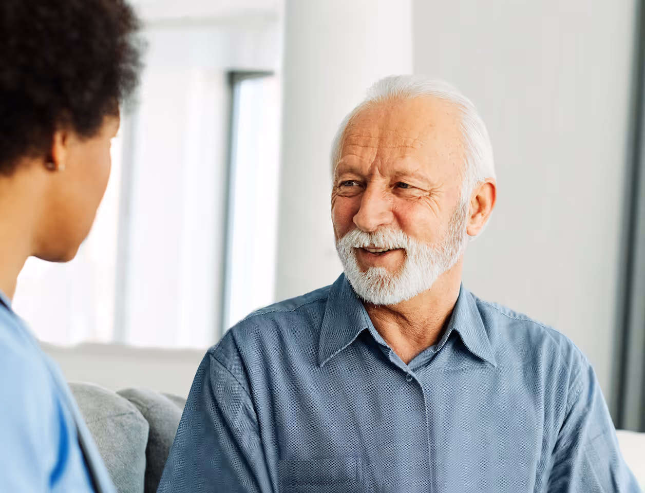 Älterer Mann mit weißem Bart und blauer Hemdbluse lächelt und spricht mit einer Krankenschwester.