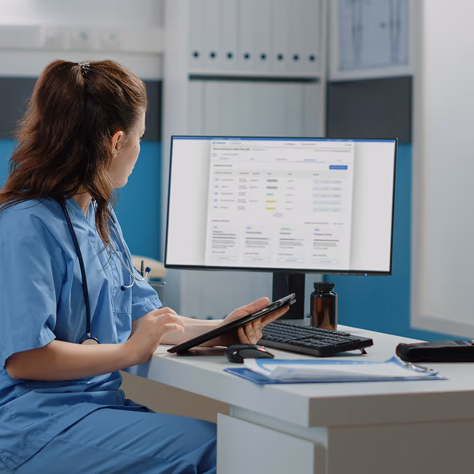 Medizinische Assistentin in blauer Uniform, die an einem Schreibtisch arbeitet und Patientendaten auf einem Tablet und Computerbildschirm überprüft.