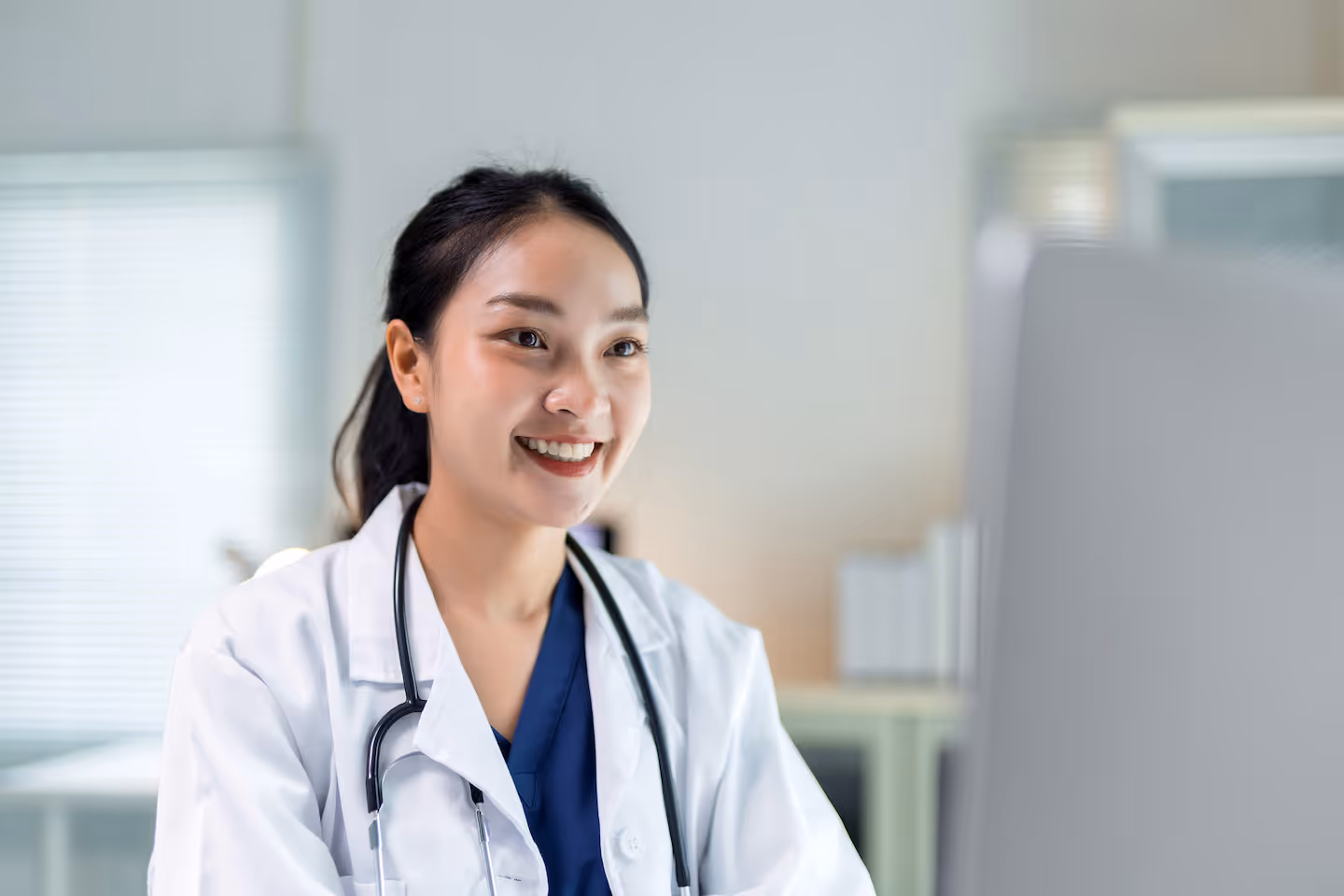 Lächelnde asiatische Ärztin mit Stethoskop vor einem Computer im hellen Büro.