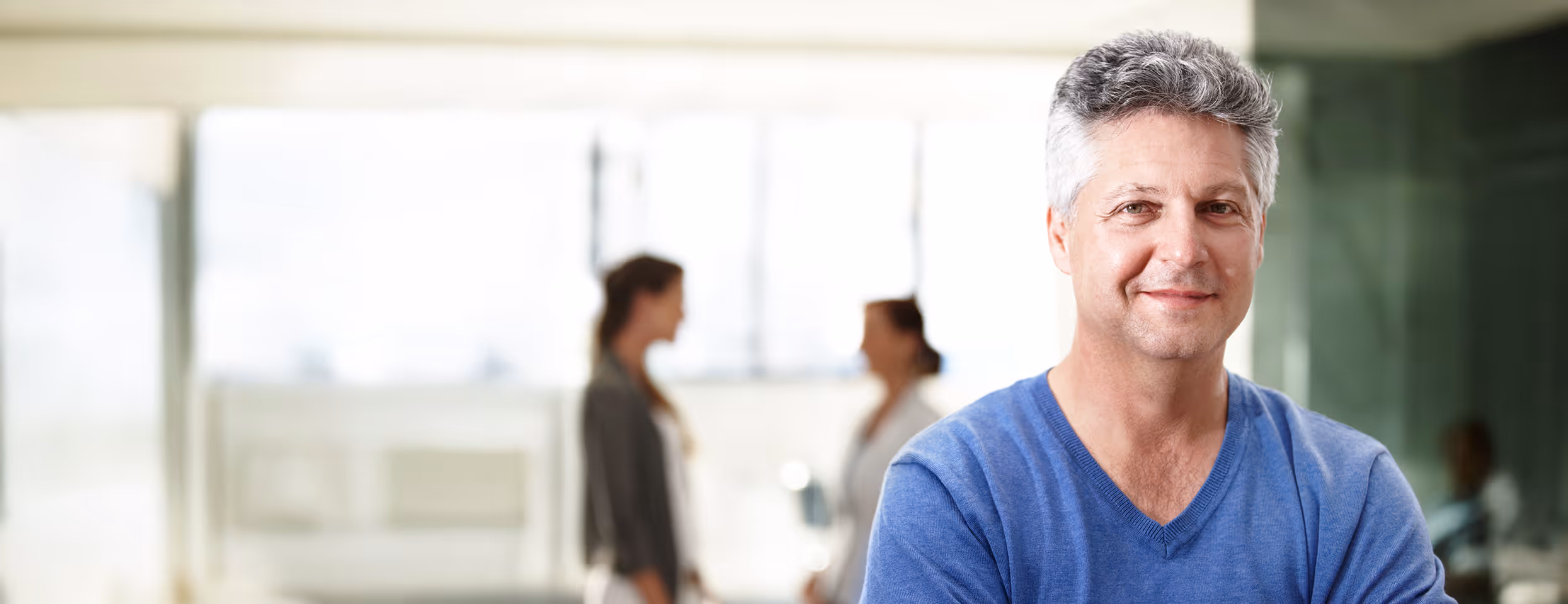 Lächelnder Mann mittleren Alters in blauem Pullover vor einem unscharfen Hintergrund mit zwei Frauen im Gespräch.